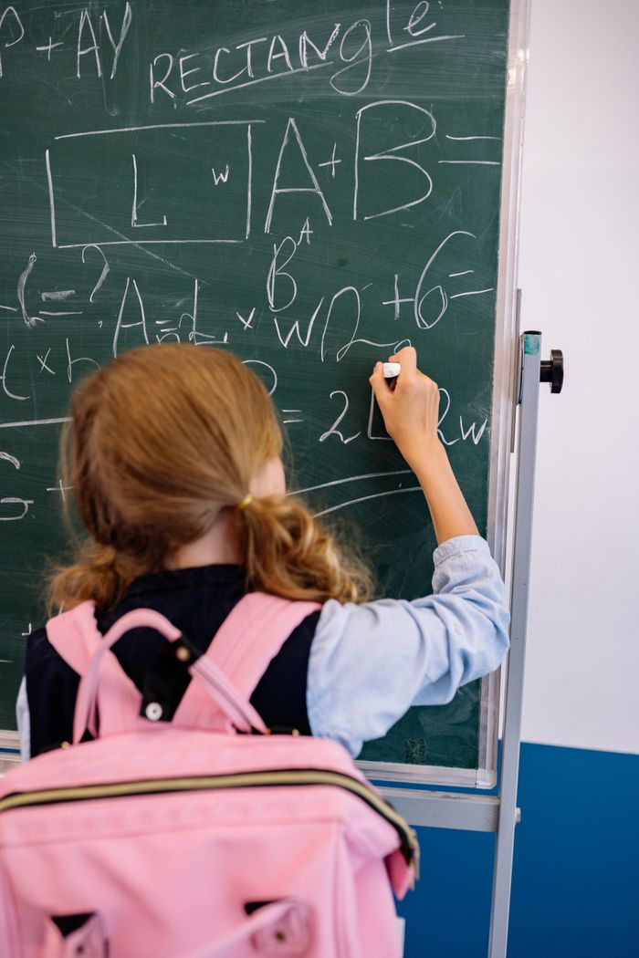 A young girl writes on a chalkboard, solving math equations in a classroom setting.