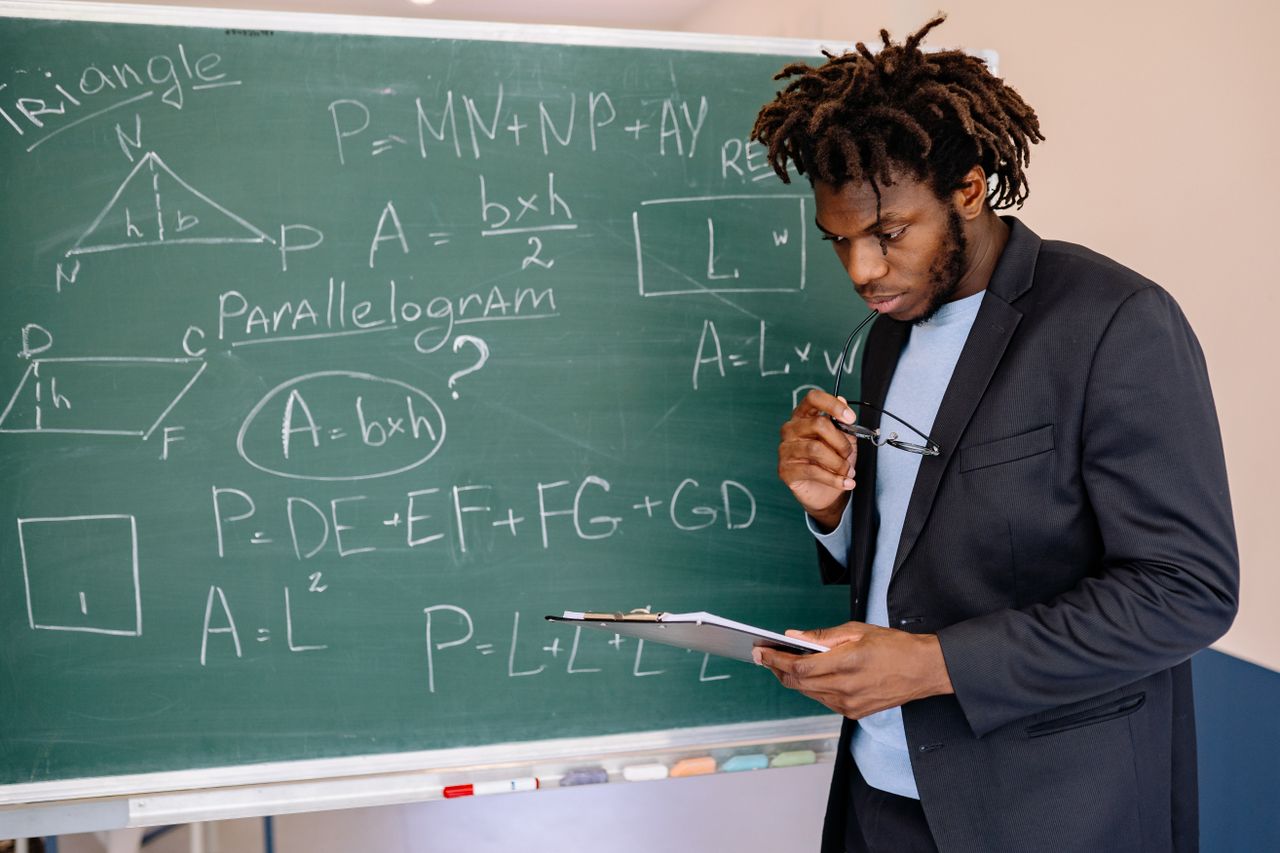 Educator in suit jacket solving math equations on blackboard, holding clipboard.